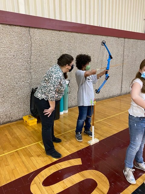 Volunteer Angie Cannon teaches a student to aim a bow and arrow in PE